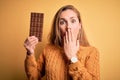 Young beautiful blonde woman holding chocolate bar standing over isolated yellow background cover mouth with hand shocked with Royalty Free Stock Photo