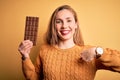 Young beautiful blonde woman holding chocolate bar standing over isolated yellow background with surprise face pointing finger to Royalty Free Stock Photo