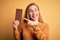 Young beautiful blonde woman holding chocolate bar standing over isolated yellow background very happy pointing with hand and Royalty Free Stock Photo