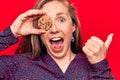 Young beautiful blonde woman holding chocolate chips cookie pointing thumb up to the side smiling happy with open mouth