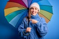 Young beautiful blonde woman holding colorful umbrella over isolated blue background very happy pointing with hand and finger Royalty Free Stock Photo