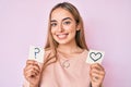 Young beautiful blonde woman holding heart and question mark reminder smiling with a happy and cool smile on face Royalty Free Stock Photo