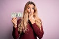 Young beautiful blonde woman holding one dollar banknote over isolated pink background cover mouth with hand shocked with shame Royalty Free Stock Photo