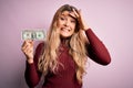 Young beautiful blonde woman holding one dollar banknote over isolated pink background stressed with hand on head, shocked with Royalty Free Stock Photo