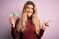 Young beautiful blonde woman holding one dollar banknote over isolated pink background very happy pointing with hand and finger to Royalty Free Stock Photo