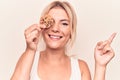 Young beautiful blonde woman holding sweet chocolate cookie over eye smiling happy pointing with hand and finger to the side