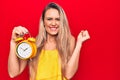 Young beautiful blonde woman holding vintage alarm clock over isolated red background screaming proud, celebrating victory and Royalty Free Stock Photo