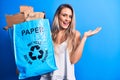 Young beautiful blonde woman recycling holding paper recycle bag full of paperboard celebrating achievement with happy smile and Royalty Free Stock Photo