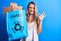 Young beautiful blonde woman recycling holding paper recycle bag full of paperboard doing ok sign with fingers, smiling friendly Royalty Free Stock Photo