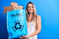 Young beautiful blonde woman recycling holding paper recycle bag full of paperboard looking positive and happy standing and Royalty Free Stock Photo