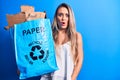 Young beautiful blonde woman recycling holding paper recycle bag full of paperboard scared and amazed with open mouth for Royalty Free Stock Photo