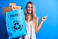 Young beautiful blonde woman recycling holding paper recycle bag full of paperboard smiling happy pointing with hand and finger to Royalty Free Stock Photo