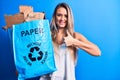 Young beautiful blonde woman recycling holding paper recycle bag full of paperboard smiling happy and positive, thumb up doing Royalty Free Stock Photo