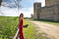 Young, beautiful, blonde woman in a red dress and flower tiara, leaning on a wooden railing next to a meadow and a castle, calm Royalty Free Stock Photo