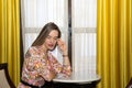 young and beautiful blonde woman sitting in an armchair next to a window covered with a curtain and yellow curtain. The woman is Royalty Free Stock Photo