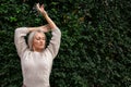 Blonde woman in sweater, stands against background of wall of green leaves, raised her hands up and closed her eyes Royalty Free Stock Photo