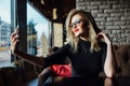 Young beautiful blonde woman taking a selfie in coffee shop. Hipster, red lips, glasses. Royalty Free Stock Photo