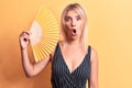 Young beautiful blonde woman using hand fan standing over isolated yellow background scared and amazed with open mouth for Royalty Free Stock Photo