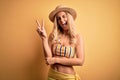 Young beautiful blonde woman on vacation wearing bikini and hat over yellow background smiling with happy face winking at the Royalty Free Stock Photo