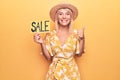 Young beautiful blonde woman on vacation wearing summer hat holding sale banner smiling happy and positive, thumb up doing Royalty Free Stock Photo