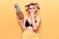 Young beautiful blonde woman wearing bikini and summer hat holding pineapple covering mouth with hand, shocked and afraid for Royalty Free Stock Photo