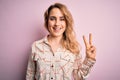 Young beautiful blonde woman wearing casual shirt standing over isolated pink background smiling with happy face winking at the Royalty Free Stock Photo