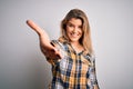 Young beautiful blonde woman wearing casual shirt standing over isolated white background smiling friendly offering handshake as Royalty Free Stock Photo