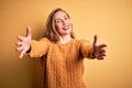 Young beautiful blonde woman wearing casual sweater standing over yellow background looking at the camera smiling with open arms Royalty Free Stock Photo