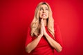 Young beautiful blonde woman wearing casual t-shirt standing over isolated red background begging and praying with hands together Royalty Free Stock Photo