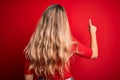 Young beautiful blonde woman wearing casual t-shirt standing over isolated red background Posing backwards pointing ahead with Royalty Free Stock Photo