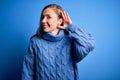 Young beautiful blonde woman wearing casual turtleneck sweater over blue background smiling with hand over ear listening an Royalty Free Stock Photo