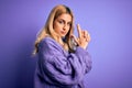 Young beautiful blonde woman wearing casual turtleneck sweater over purple background Holding symbolic gun with hand gesture, Royalty Free Stock Photo