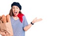 Young beautiful blonde woman wearing french beret holding bag with bread celebrating victory with happy smile and winner