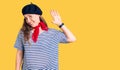 Young beautiful blonde woman wearing french beret and striped t-shirt waiving saying hello happy and smiling, friendly welcome