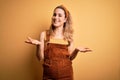Young beautiful blonde woman wearing overalls and diadem standing over yellow background smiling cheerful with open arms as Royalty Free Stock Photo