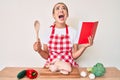 Young beautiful blonde woman wearing professional baker apron reading cooking recipe book angry and mad screaming frustrated and Royalty Free Stock Photo