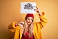 Young beautiful blonde woman wearing raincoat and wool cap holding banner with cloud image with angry face, negative sign showing