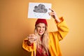 Young beautiful blonde woman wearing raincoat and wool cap holding banner with cloud image annoyed and frustrated shouting with