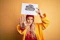 Young beautiful blonde woman wearing raincoat and wool cap holding banner with cloud image with open hand doing stop sign with