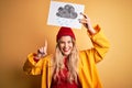 Young beautiful blonde woman wearing raincoat and wool cap holding banner with cloud image surprised with an idea or question