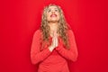 Young beautiful blonde woman wearing red casual t-shirt standing over isolated background begging and praying with hands together Royalty Free Stock Photo