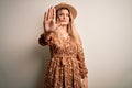 Young beautiful blonde woman wearing summer dress and hat over isolated white background doing stop sing with palm of the hand Royalty Free Stock Photo