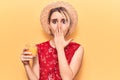 Young beautiful blonde woman wearing summer hat drinking glass of orange juice covering mouth with hand, shocked and afraid for Royalty Free Stock Photo