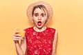 Young beautiful blonde woman wearing summer hat drinking glass of orange juice scared and amazed with open mouth for surprise, Royalty Free Stock Photo