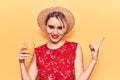 Young beautiful blonde woman wearing summer hat drinking glass of orange juice smiling happy pointing with hand and finger to the Royalty Free Stock Photo