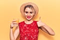 Young beautiful blonde woman wearing summer hat drinking glass of orange juice smiling happy and positive, thumb up doing Royalty Free Stock Photo