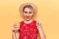 Young beautiful blonde woman wearing summer hat drinking glass of orange juice smiling with an idea or question pointing finger Royalty Free Stock Photo