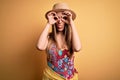 Young beautiful blonde woman wearing swimsuit and summer hat over yellow background doing ok gesture like binoculars sticking Royalty Free Stock Photo