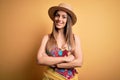 Young beautiful blonde woman wearing swimsuit and summer hat over yellow background happy face smiling with crossed arms looking Royalty Free Stock Photo