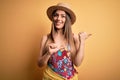 Young beautiful blonde woman wearing swimsuit and summer hat over yellow background Pointing to the back behind with hand and Royalty Free Stock Photo